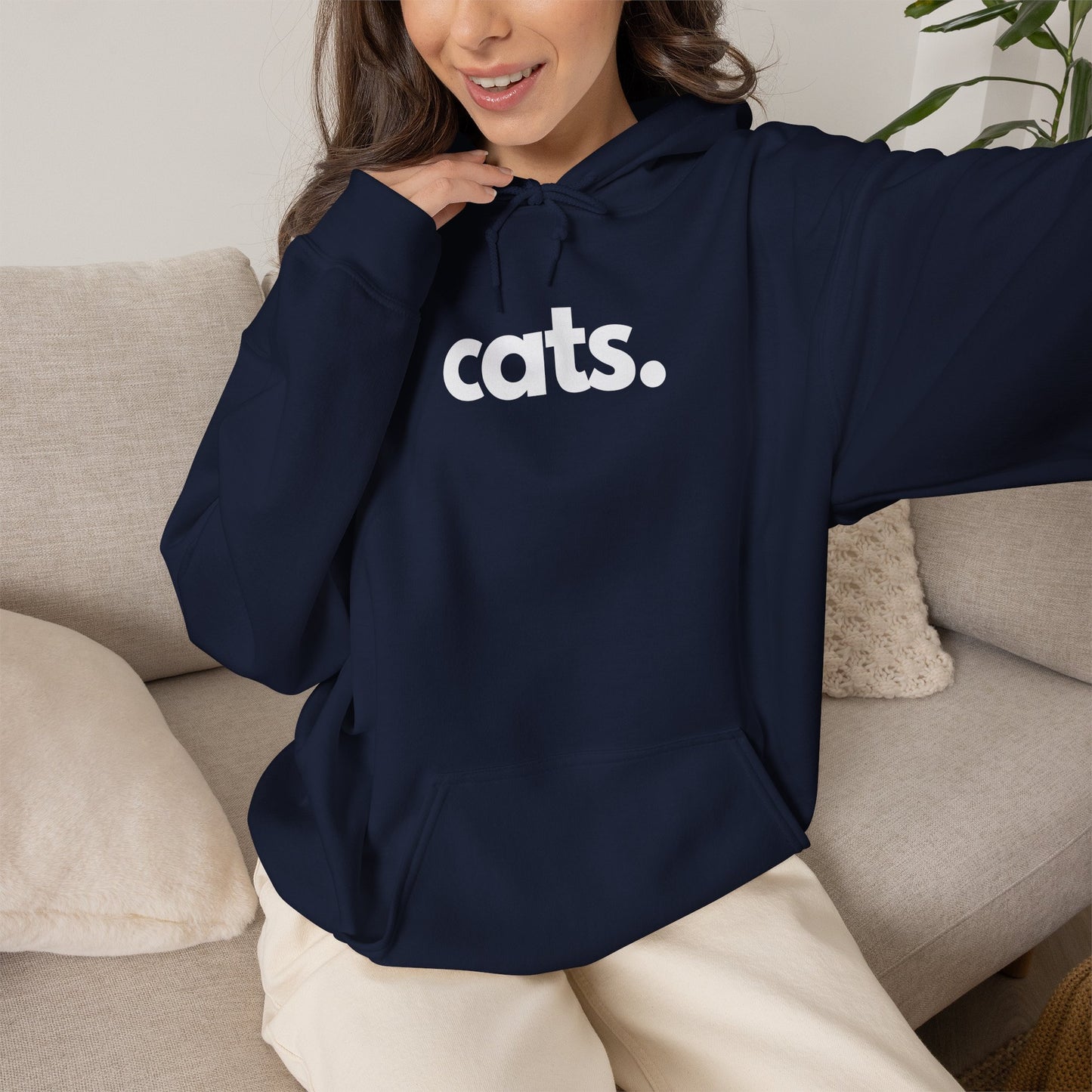 A woman wearing a navy blue hoodie with the word 'cats.' printed in white across the chest, sitting on a light beige couch.
