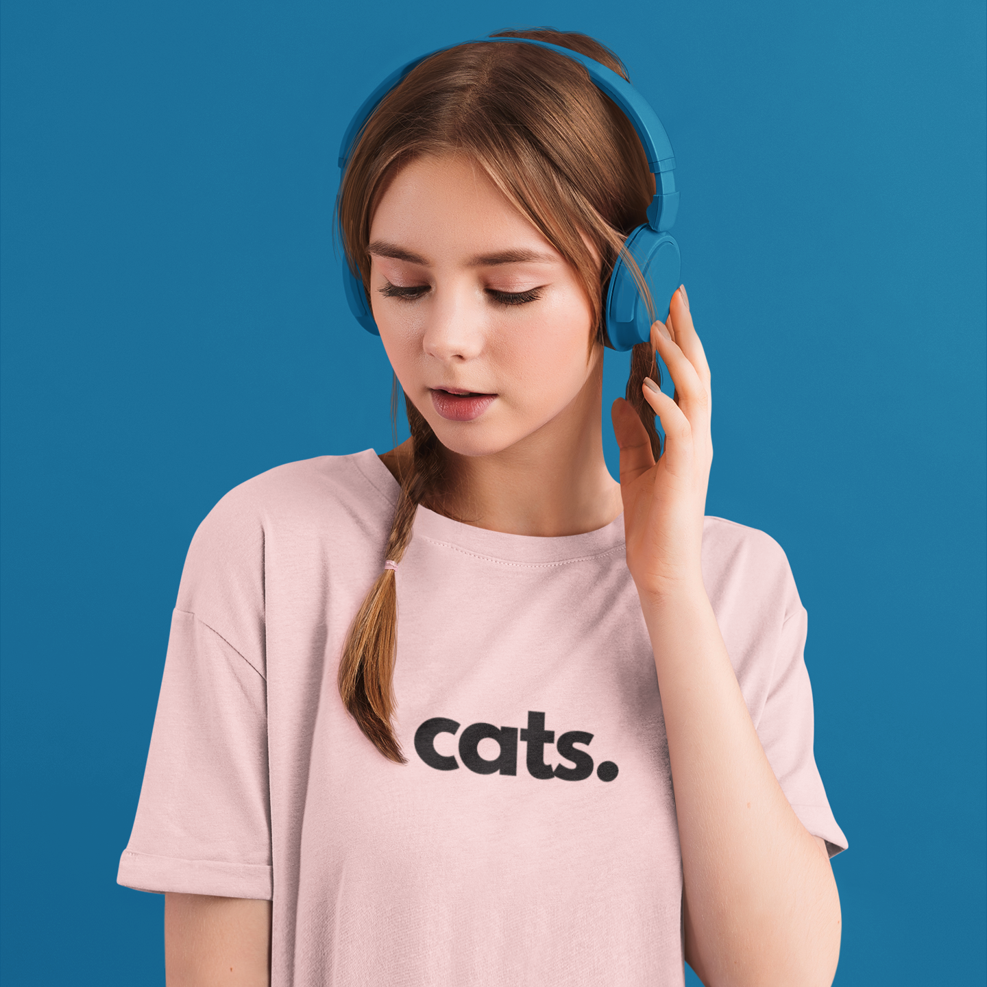 A young woman with brown hair in a side braid wears bright blue over-ear headphones and a light pink t-shirt with the word 'cats.' printed in black across the chest, set against a solid blue background.